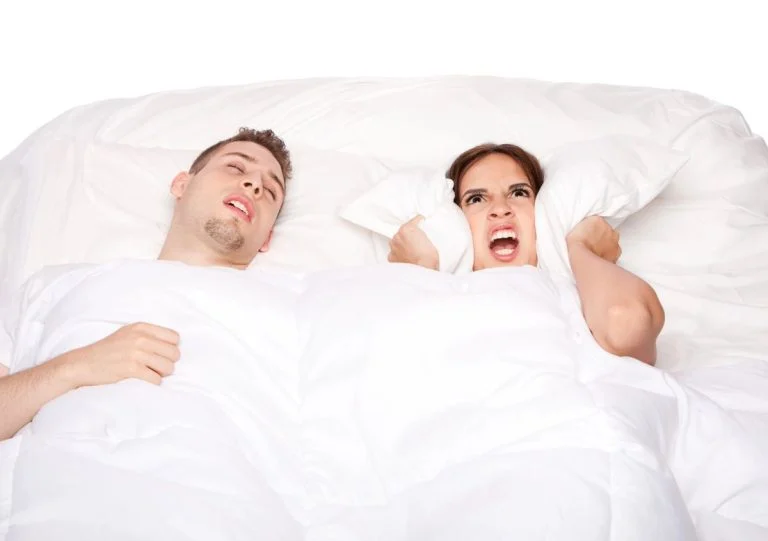 Frustrated woman holding a pillow over her ears as a man snores in bed